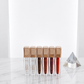 Miss Rose Lip Void liquid lip gloss 6-piece collection lined up in rose gold square bottles on white marble surface with crystal prism showing clear, nude, chocolate brown, berry, deep burgundy and red shades