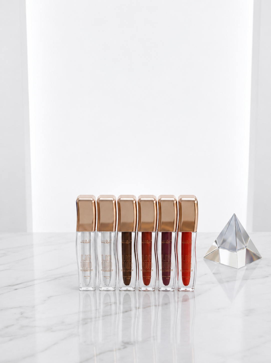 Miss Rose Lip Void liquid lip gloss 6-piece collection lined up in rose gold square bottles on white marble surface with crystal prism showing clear, nude, chocolate brown, berry, deep burgundy and red shades
